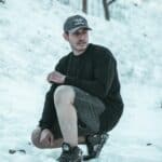Stylish young man in winter outdoor attire kneeling on snow, with trees and a winter landscape behind.
