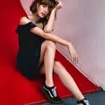 Fashion model wearing black lace dress and sneakers, sitting on a colorful floor with pink and red wall background.